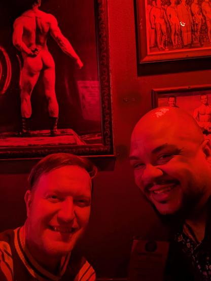 Me and Billy at a bar table under red lighting with a framed pic of a muscular man's butt hanging on the wall