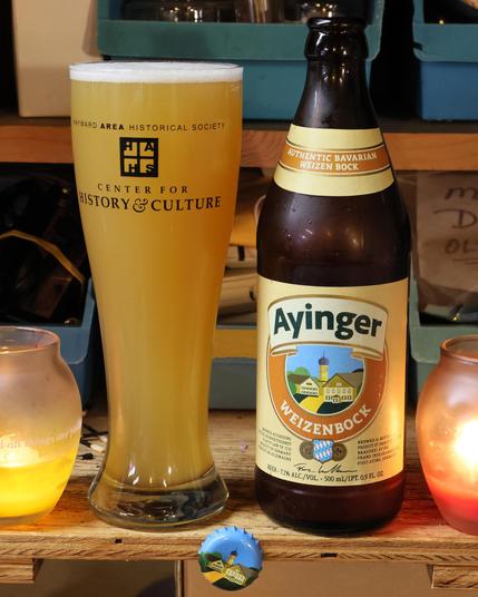 A pilsner glass filled with a straw-colored hazy liquid and topped with a white layer of foam stands next to a beer bottle. The label is light yellow, gold, orange, and green, and reads “Ayinger Weizenbock.”