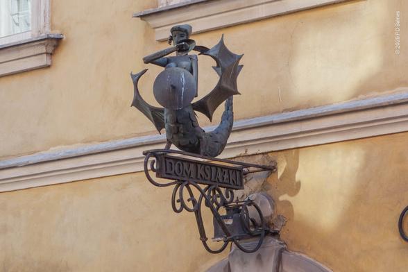 The heraldic symbol for the city of Warsaw rendered in beaten metal, the Warsaw mermaid or melusina holding a sword and shield, above a wrought iron sign for a bookshop