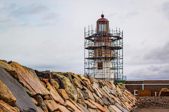 Photo of a white lighthouse with a copper lantern room surrounded by scaffolding. A granite jetty is in the foreground.