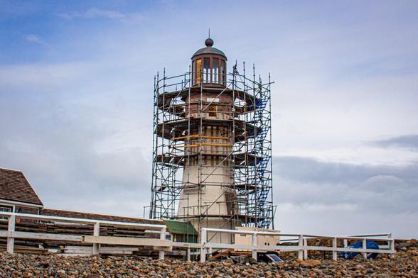 Photo of a white lighthouse with a copper lantern room surrounded by scaffolding.