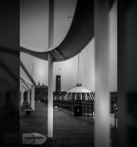 Monochrome shot of harbour walkway, with curved raised feature over benches and covered circular viewing area in background