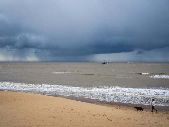 A wide, eye-level shot depicts a sandy beach, a body of water, and a stormy sky. In the distance, a fishing boat and a red boat are visible on the water. A person is walking a black dog on the sand near the water's edge. The sky is covered with dark, ominous clouds, and rain is falling in the distance. The water is choppy, with small waves breaking on the shore. The sand is light brown and extends into the foreground.