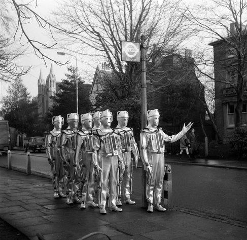 a group of cybermen from Dr Who waiting for a bus, one has its arm raised as if to imply "I remember when this was all trees"
