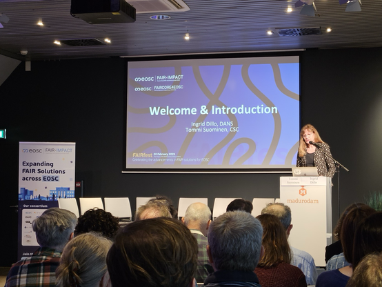 Phot of a stage with the speaker (Ingrid Dillo from DANS) standing at a podium in front of a projection screen with a blue slide that reads welcome & introduction. On the left, a white roll up banner of FAIR IMPACT.
