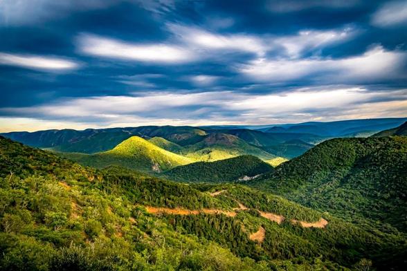 The image depicts a scenic view of a landscape with lush green mountains and valleys under a dramatic sky. The clouds are moving, creating patches of sunlight across the hills. The overall atmosphere is serene and picturesque.