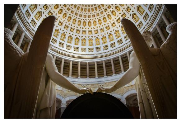 Zwei große, sakrale Engelstatuen stehen nebeneinander in einem gewölbten, kreisförmigen Raum. Die Engel halten sich an den Händen und blicken zu einer kunstvoll gestalteten Decke hinauf. Der Kontext umfasst ein monumentales, historisches Gebäude mit Säulen und einem beeindruckenden Deckendekor.