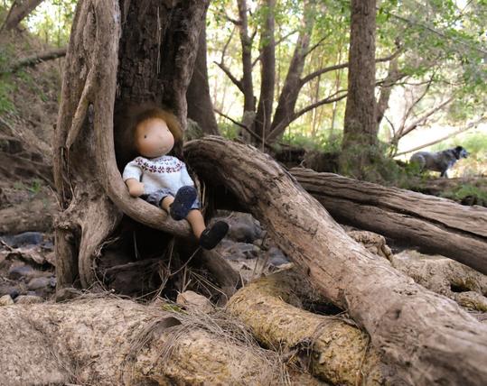 A waldorf-inspired doll wearing a Fair Isle jumper is sitting in the bush by a creek