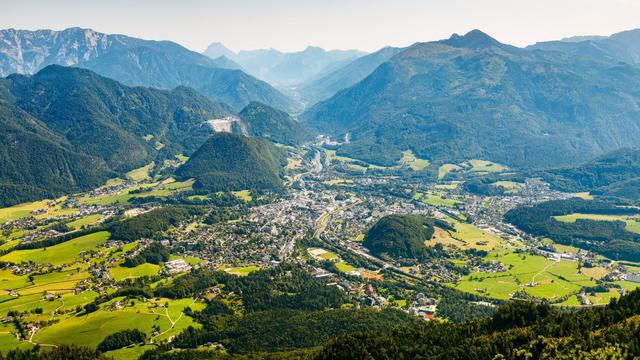 Das Bild zeigt eine malerische Landschaft mit einer kleinen Stadt in einem Tal, umgeben von grünen Hügeln und hohen Bergen. Der Blickwinkel ist von einem hohen Berg auf das Tal hinab. Die Gegend wirkt sehr idyllisch, mit vielen Wiesen, Wäldern und verstreuten Häusern. Eine Fluss schlängelt sich durch das Tal und die Stadt. Die Berge im Hintergrund verleihen dem Bild eine beeindruckende Tiefe.