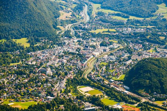 Das Bild zeigt eine Aufnahme von Bad Ischl von schräg oben.
Zentrum der Stadt: Deutlich erkennbar ist das Stadtzentrum mit seinen vielen historischen Gebäuden, engen Straßen und einer dichten Bebauung. Die Architektur wirkt typisch österreichisch, mit charmanten Fassaden und klassischen Strukturen.
Fluss: Ein Fluss (die Traun) schlängelt sich durch die Stadt und wird von mehreren Brücken überquert. Dies verleiht der Stadt ein malerisches Flair.
Umgebung: Die Stadt ist von grünen Hügeln und bewaldeten Bergen umgeben, die die Landschaft besonders idyllisch machen.
Verkehrswege: Eine Bahnstrecke und Straßen sind erkennbar, die Bad Ischl mit anderen Orten verbinden.
Freiflächen und Parks: In der Umgebung gibt es viele grüne Wiesen, Gärten und Sportplätze, die zur Erholung einladen.