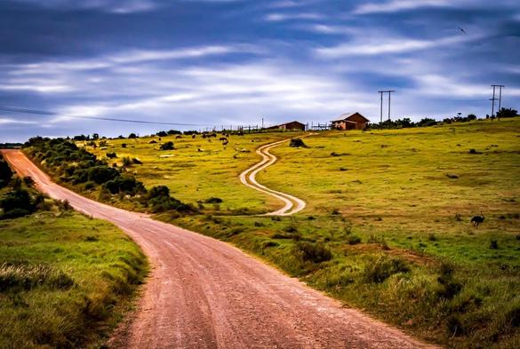 The image features a rural landscape with a dirt road winding through grassy fields. There are hills in the background, topped with a structure that looks like a house or barn. Power lines are visible, and the sky above is cloudy with a slightly dramatic appearance. The area appears open and expansive.