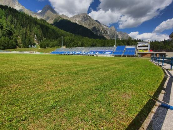 In this picture you can see a grass sportsfield and a stand with mostly blue plastic seats in front of the forest and the mountains. Some seats are white and form the name "Antholz". The sky is blue but covered with some grey clouds.