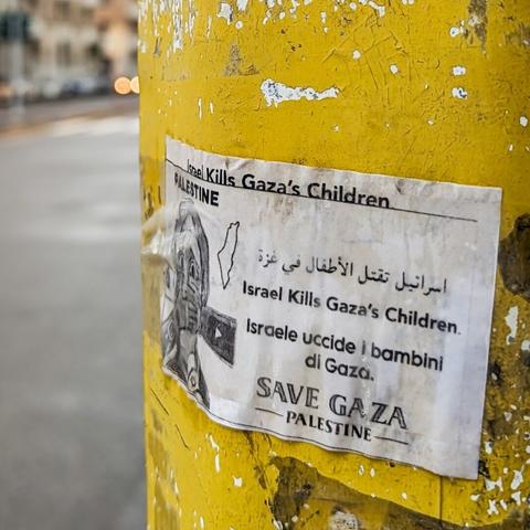 A close up of a sticker on a yellow light post that reads in a few languages "Israel kills Gaza's children. Save Gaza "
