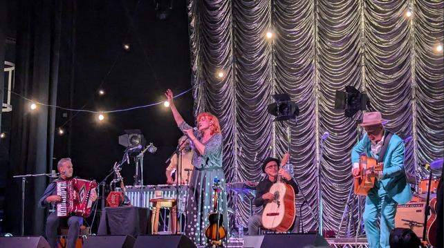 A picture of he stage at a concert, the female singer standing in the centre holding the microphone stand in one hand, her other arm raised in the air.
An accordion player is sitting on the left. a man playing the marimba is partially hidden behind the singer, sitting at the back is a man playing an acoustic bass guitar, and the lead acoustic guitar player in on the right of the image. The drummer is just off to the right of the image.
Monitor speakers can be seen at the front of the stage, and there's a black and silver backdrop behind the band