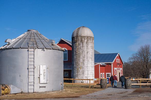 A rustic farm scene with a red barn, a weathered concrete silo, and a metal grain bin under a clear blue sky. Three people walk toward the barn on a gravel path, passing wooden barrels and a wooden fence.