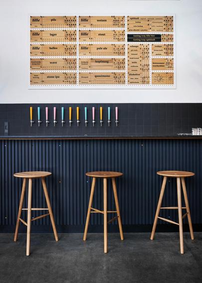 Three bar stools in front of a counter. On the wall behind them are wooden boards on which various types of beer are labelled.