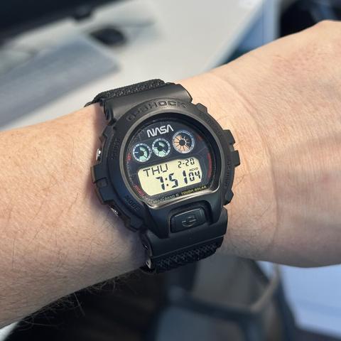 Round digital watch with black resin case cover and bezel, two pushers on each side, digital display in bottom of the black screen showing day of the week, month and day, and hours-minutes-seconds. Three round digital subdials in the top half of the screen: two smaller ones with blue-green background showing state of charge and active functions, the larger one with pinkish-red backgound showing a seconds countdown that repeats every ten seconds. The NASA "worm" logo is at the very top of the screen.