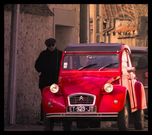 La photographie représente une Citroën 2CV rouge vif garée dans une rue. À côté de la voiture, une personne portant des vêtements sombres et une casquette se tient dans l'ombre, sur le trottoir. La scène semble se dérouler dans un cadre pittoresque, périurbain.