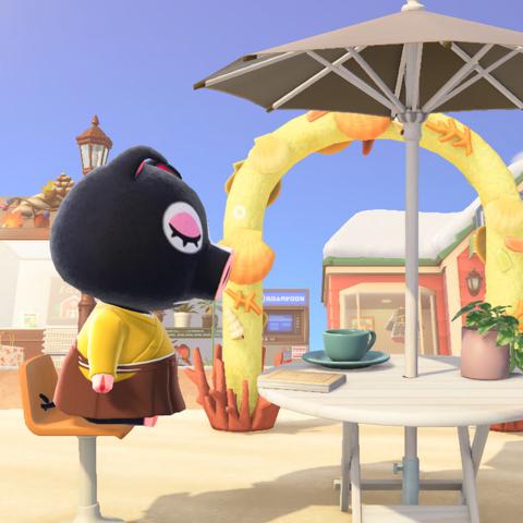 Animal Crossing: New Horizons screenshot. Image shows Agnes (black pig) sleeping while sitting at a table with an umbrella on the beach with a book / coffee cup right in front of her. Shops are visible in the background behind her.