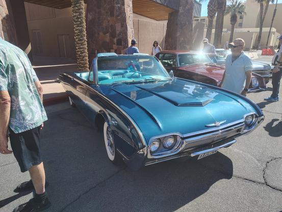 A dark turquoise 1961 Ford Thunderbird convertible is backed into a parking space at the Palm Springs, California convention center for a classic car show.  The photo is from eye height, three quarters view from the front passenger's side.  The top is down, revealing a light turquoise bucket seat interior.  Wire wheels are visible, with wide whitewall tires.
