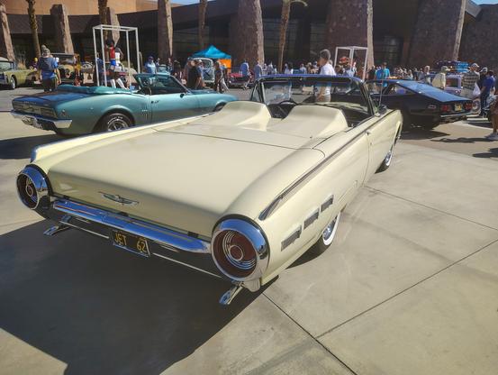 A pale yellow 1962 Thunderbird convertible is seen with its top down from a low angle on the passenger's side from the rear, highlighting the very long sweep of the trunk and the fiberglass tonneau cover.  The tonneau hides the back seats and has molded headrests behind the driver and front passenger's seats.  

Visible behind the Thunderbird is a first-generation Pontiac Firebird 400 convertible in aqua. A Ferrari is parked in front of the Thunderbird.