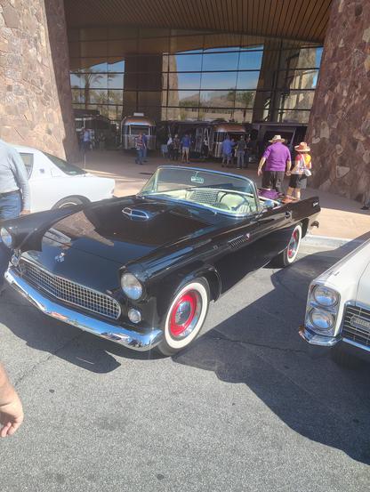 A black 1955 or 1956 first-generation Thunderbird roadster was on display at the classic car show, seen here from the front. It has red steel wheels with chrome trim rings and hubcaps, and a white vinyl bench seat interior.  A black and white houndstooth lap rug is draped across the back of the passengers seat.  Just visible in the picture are a white Studebaker Avanti to the left and a cream mid-60s Cadillac to the right.