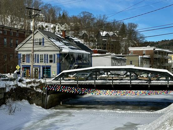 A frozen river and an old iron bridge with an art installation across the frozen river that looks like colorful bunting.