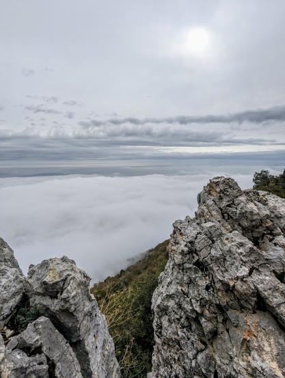 A rocky outcrop with sparse vegetation overlooks a dense layer of fog filling the Lake Garda valley under a cloudy sky.
