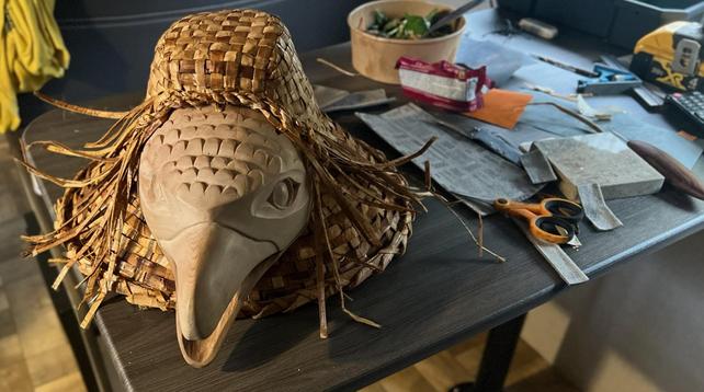 A cedar raven head carving attached to a woven cedar hat is on a table. Around it are scattered tools & materials, including scissors, a file, sandpaper, a wooden bowl & some colorful papers.