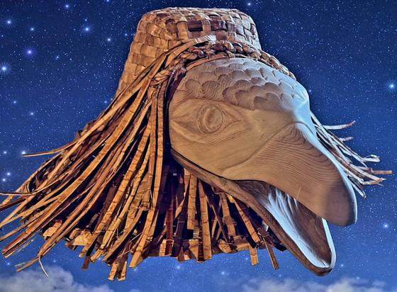 Low-angle close-up shot of a Coast Salish Indigenous cedar wood carving against a deep blue night skies background. The subject is a detailed raven's head & face. The raven is tan with a visible wood grain texture. It is attached to a cedar woven hat with some woven fiber fringe that drapes around the raven's head. The blue background includes a faint layer of white clouds right above the horizon. Sprinkled throughout the blue is an effect of twinkling stars in different sizes.