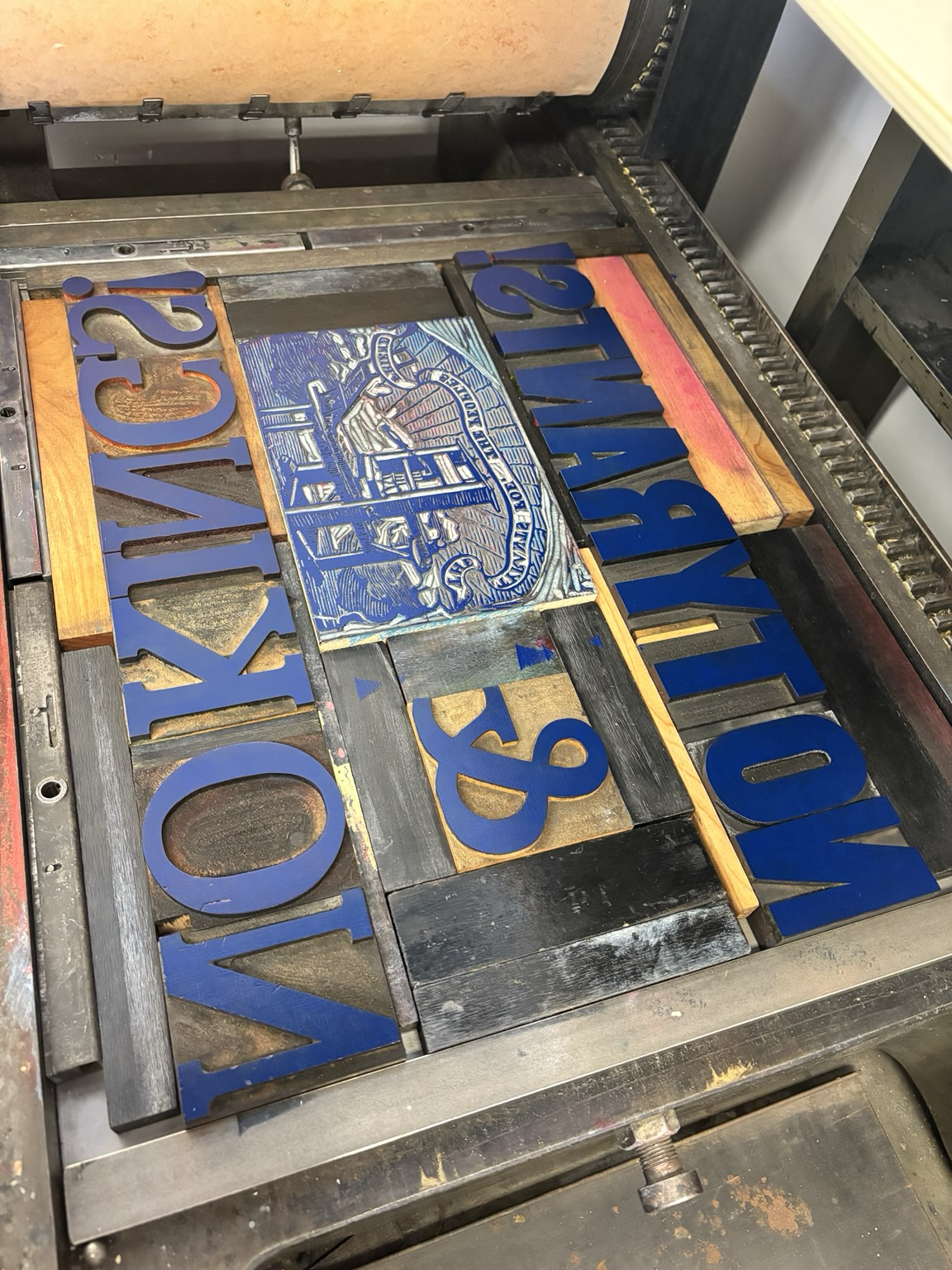 A printing press bed with wood type inked with blue ink reading:  NO TYRANTS! & NO KINGS!   In large wood type font. There is a large image cut of a wooden printing press with a banner of text above it reading “THE TYRANTS FOE THE PEOPLES FRIEND”