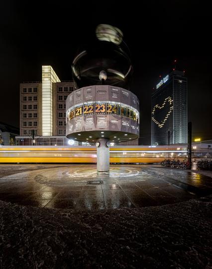 The image captures the World Clock at Alexanderplatz in Berlin at night. Due to a long exposure, the rotating upper part of the clock appears blurred. A yellow tram passes in the background, leaving a horizontal light trail. To the right, the Park Inn Hotel is visible, with its windows forming the shape of a heart. The wet pavement reflects the artificial lights of the surroundings, creating an atmospheric scene.