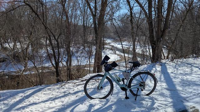 Sage green Vado SL ebike parked off the Zumbro River South Trail with the partially open river behind in a wooded area with snow on the ground in bright sunshine.