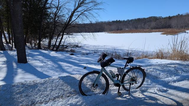 Sage green Vado SL ebike parked off Mayowood Road with the snow covered and frozen Mayowood Lake behind in a wooded area with snow on the ground in bright sunshine.