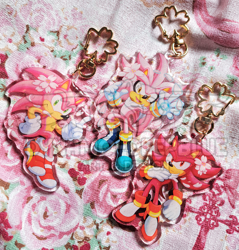 A photograph of glitter acrylic charms depicting Sonic, Silver and Shadow with pink fur and cherry blossoms. Each charm has a gold cherry blossom-shaped clasps. The charms are placed on top of a pink dress with a rose pattern for the backdrop.