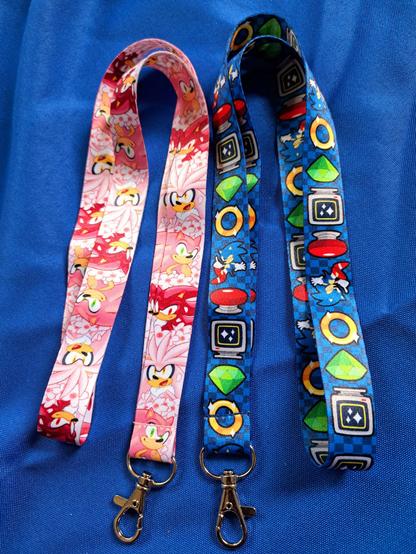 A photograph of two Sonic-inspired lanyards. One features Sonic, Shadow and Silver with pink fur and cherry blossoms in a repeating pattern. The other lanyard features a repeating pattern depicting Classic Sonic, a red spring, an invincibility monitor, a green Chaos Emerald, and a ring, against a blue checkered background.