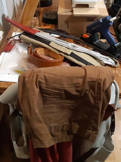 Clothes and props and wig of a cosplay over a stool and on a messy table, sewing machine and hot glue gun in the background
