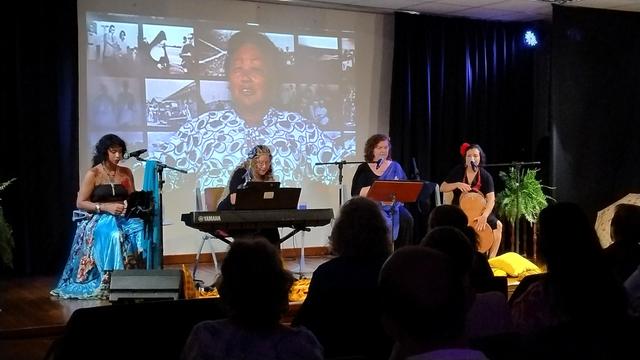 No palco há quatro mulheres se apresentando em um palco na Biblioteca Demonstrativa de Brasilia. Ao fundo passa um video de diversas entrevistas com pioneiras