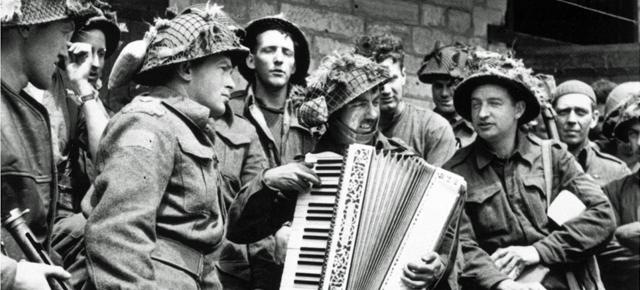 A group of combat-ready WWII-era Canadian soldiers gather around one of their number serenading them on a piano accordion.