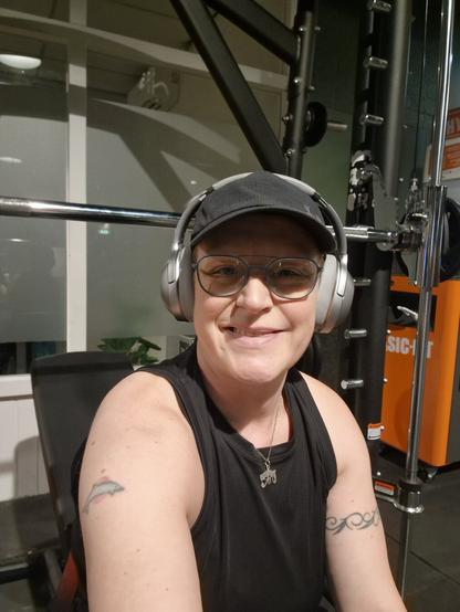 Selfie of a person in the gym. Sitting on an incline bench, surrounded by the Smith Machine. Smiling at the camera. Wearing a black top, black cap, silver headphones and blue-rimmed glasses.