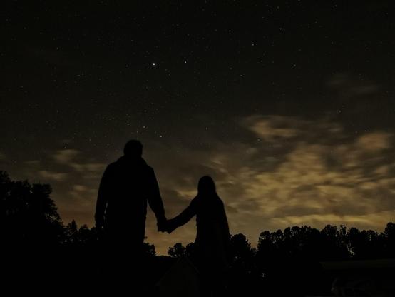 In the foreground, the black silhouettes of father and daughter hold hands. In the background you see the night sky. The trees are silhouetted on the bottom of the frame. Above them glow some clouds by the light of a nearby city. The top half of the frame features the stars and constellations. Sirius shines bright