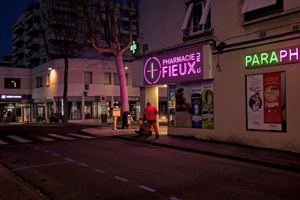 A crossroads in the city at night. The wall and sign of a pharmacy in front of which a cleaning agent in orange clothes passes. On the other side of the intersection a bank ATM with a silhouette in front of it. The nocturnal atmosphere is in the fluorescent orange/pink/green colors.