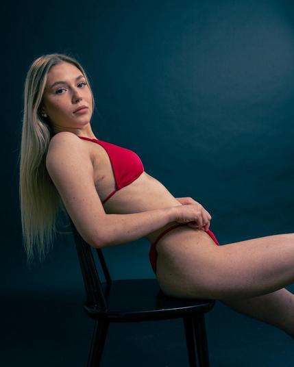 The image shows a person with long blonde hair wearing a red bikini, sitting on a black chair. The background is a dark color.