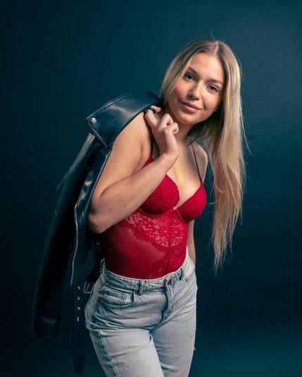 The image shows a person posing against a dark background. They are wearing a red lace top and light-colored jeans, with a black leather jacket draped over one shoulder. The individual has long, blonde hair and is smiling slightly at the camera.