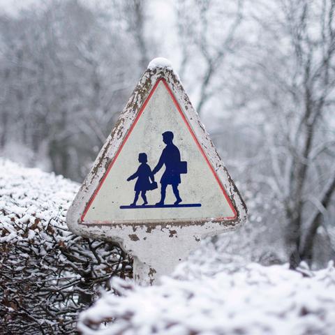 L'image montre un panneau de signalisation triangulaire indiquant un passage pour écoliers. Le panneau, bordé de rouge, présente les silhouettes d'un adulte tenant la main d'un enfant. Il est usé et partiellement recouvert de neige, notamment sur son sommet. À l'arrière-plan, des arbres dénudés et des buissons sont recouverts d'une fine couche de neige, créant une ambiance hivernale calme et brumeuse.