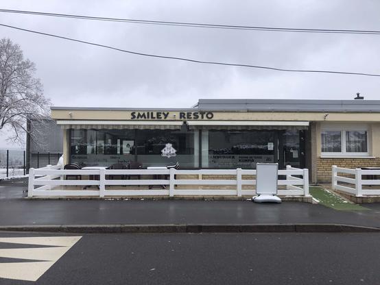 L'image montre la façade d'un restaurant nommé "SMILEY - RESTO" par temps gris et froid. Il ressemble au Diner américain ouvert 24/24. Devant l'établissement, une terrasse clôturée par une barrière blanche contient plusieurs chaises en plastique marron disposées autour de tables. Un léger manteau de neige recouvre le sol et certaines parties du paysage en arrière-plan.