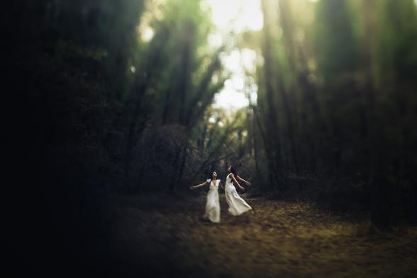 Photographie de deux femmes en robes blanches dansant dans une forêt. Le flou général donne une atmosphère onirique.