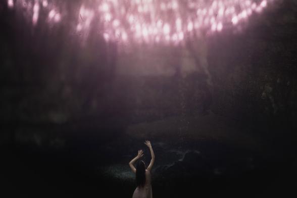 Photographie en freelensing d'une femme de dos qui lève les bras dans un geste de protection. Elle est dans une cavité sombre, est habillée d'une robe rose. Un artefact de lumière, rose lui aussi, surgit du haut de la photo.