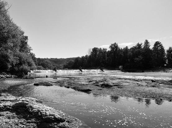 Ein ruhiger Fluss fliesst durch eine felsige Landschaft, umgeben von Bäumen und einem kleinen Wasserfall im Hintergrund.