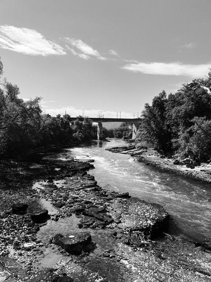 Ein Fluss fliesst durch eine felsige Landschaft mit einer Brücke im Hintergrund, umgeben von Bäumen und einem klaren Himmel.